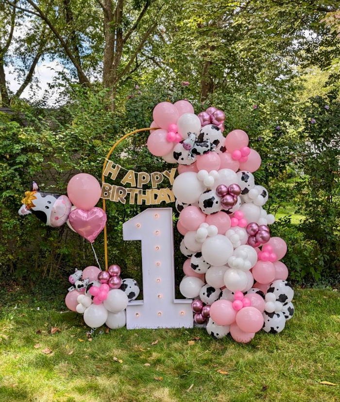 Decorations for a first birthday celebration with pink, white, and cow-patterned balloons, a large white number 1, and a gold banner that says 'Happy Birthday' on a yellow arch, set outdoors among trees and bushes.