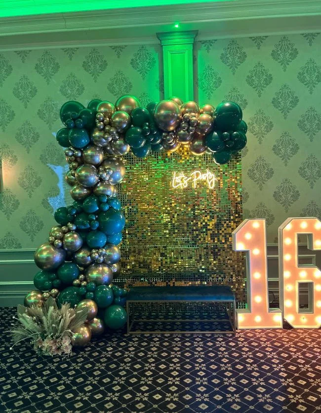 Decorative party setup with a balloon arch in shades of green, gold, and silver, a sequin backdrop with a neon sign that says "Let's Party."
