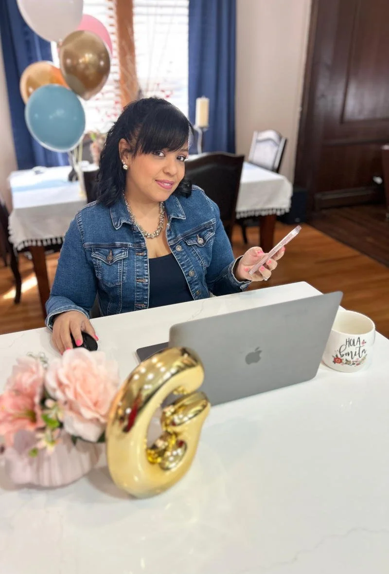 A woman in a denim jacket sitting at a table with a laptop and phone.