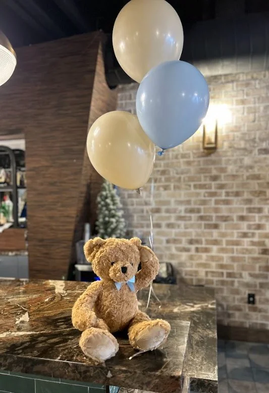 A teddy bear with a blue bow tie sitting on a marble countertop holding three balloons.