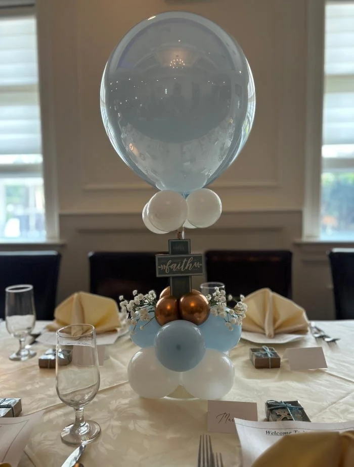 Elegant table centerpiece featuring a large clear balloon, decorated with white, blue, and gold smaller balloons, and surrounded by small flowers, placed on a white tablecloth with champagne glasses, napkins, and small gifts for a celebration.