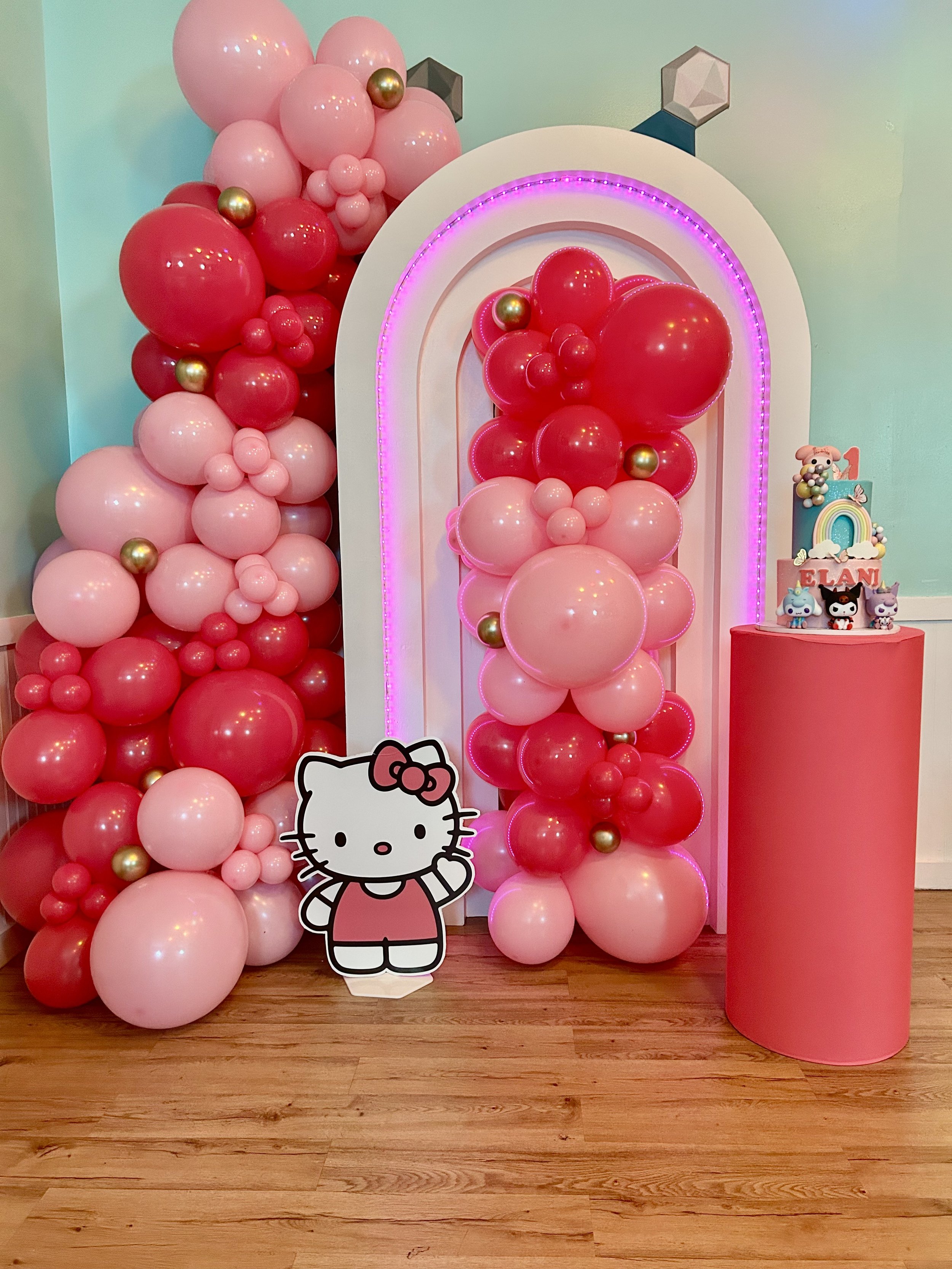Decorative birthday setup with pink and red balloons, a Hello Kitty cut-out, a colorful cake with character toppers, and a pink cylindrical stand, all on a wooden floor against a light green wall.