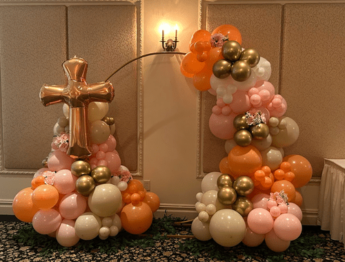 Decorative balloon arrangements with pink, orange, white, gold, and silver balloons, shaped with a cross.