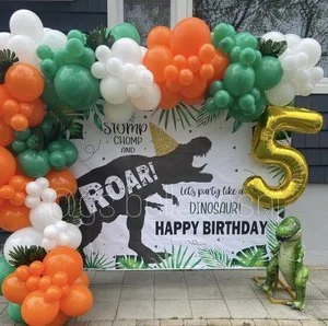 Colorful balloon arch in green, white, orange, and gold at a birthday party with a dinosaur-themed backdrop saying 'Roar! Let's party like a dinosaur! Happy birthday' and a large gold number 5 balloon.
