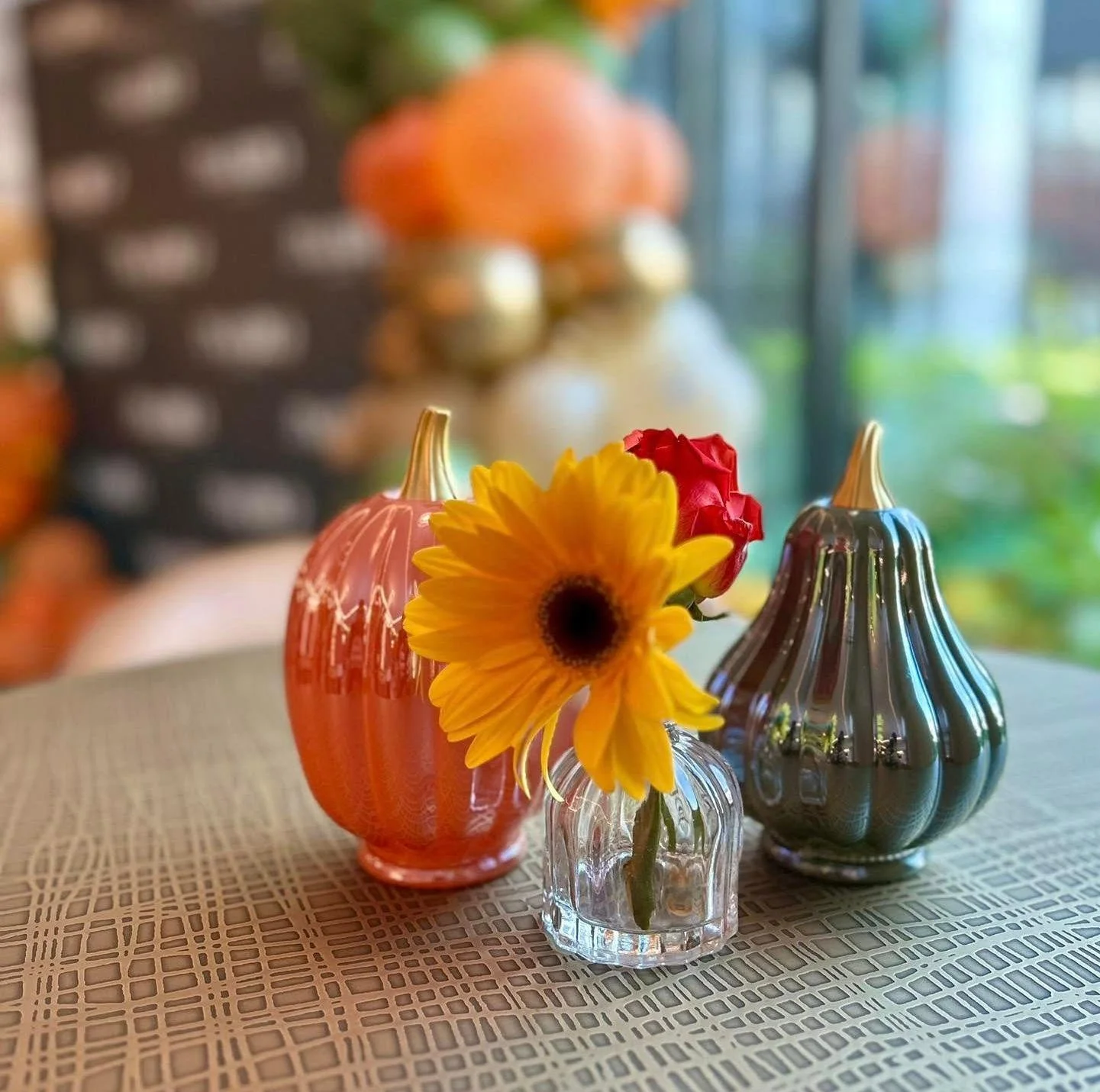 Three small decorative vases, one pink, one metallic silver, and one clear, with flowers including a yellow sunflower, a red rose, and a small red flower, on a textured tablecloth.