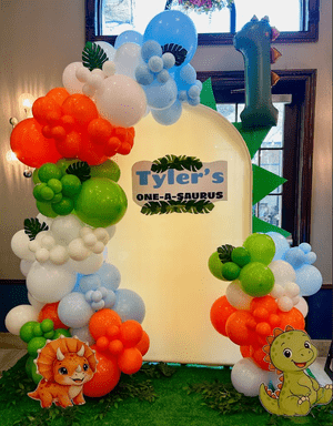 Colorful balloon arch with dinosaurs and cake decorations marking a first birthday celebration.