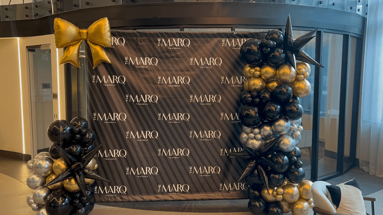 Decoration with gold, black, and silver balloons around a step-and-repeat backdrop featuring 'The MARQ' logo, with a large gold bow on the top left corner.