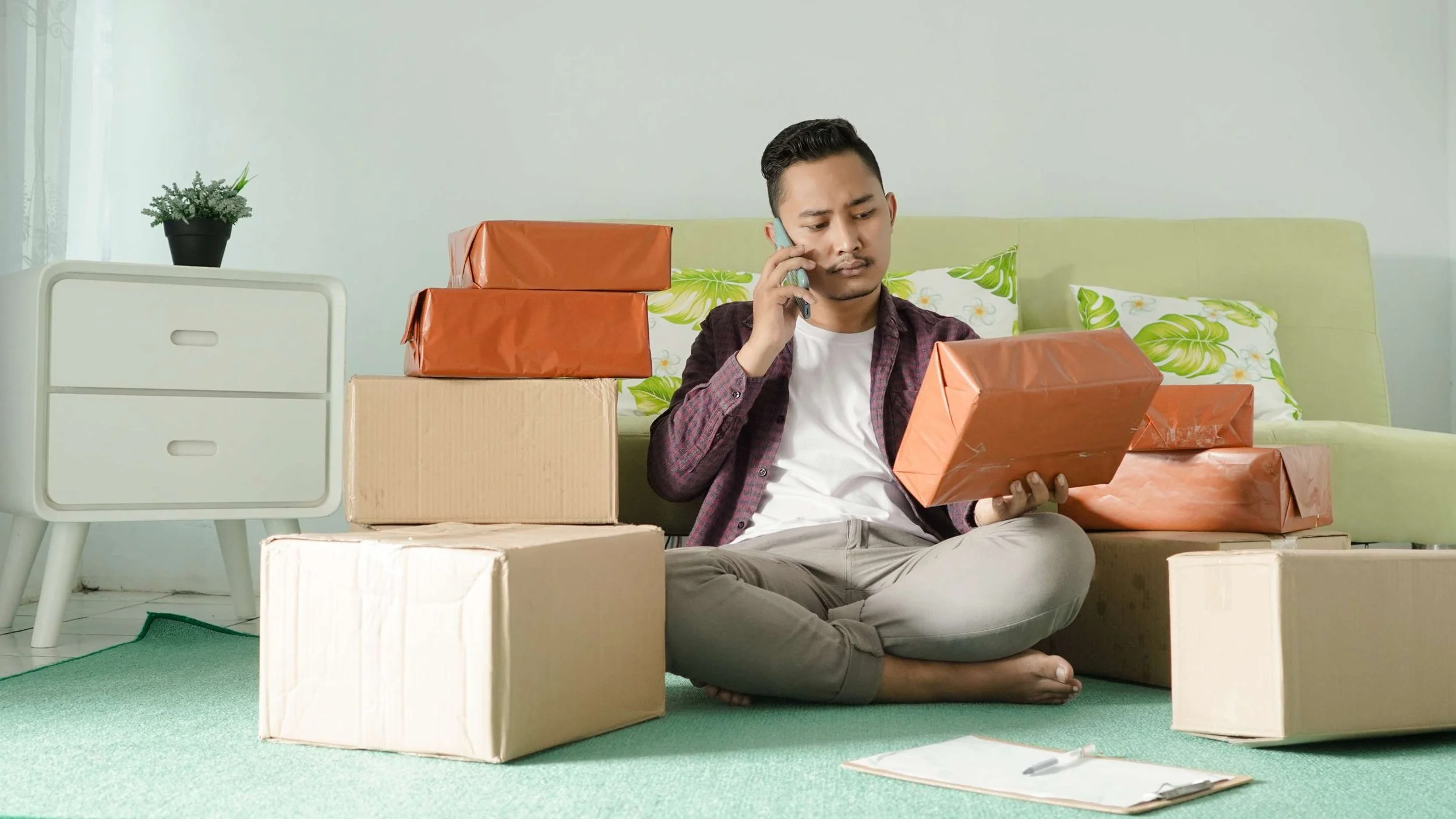 asian man sitting on the ground by his couch looking at a package wrapped in orange wrapping paper