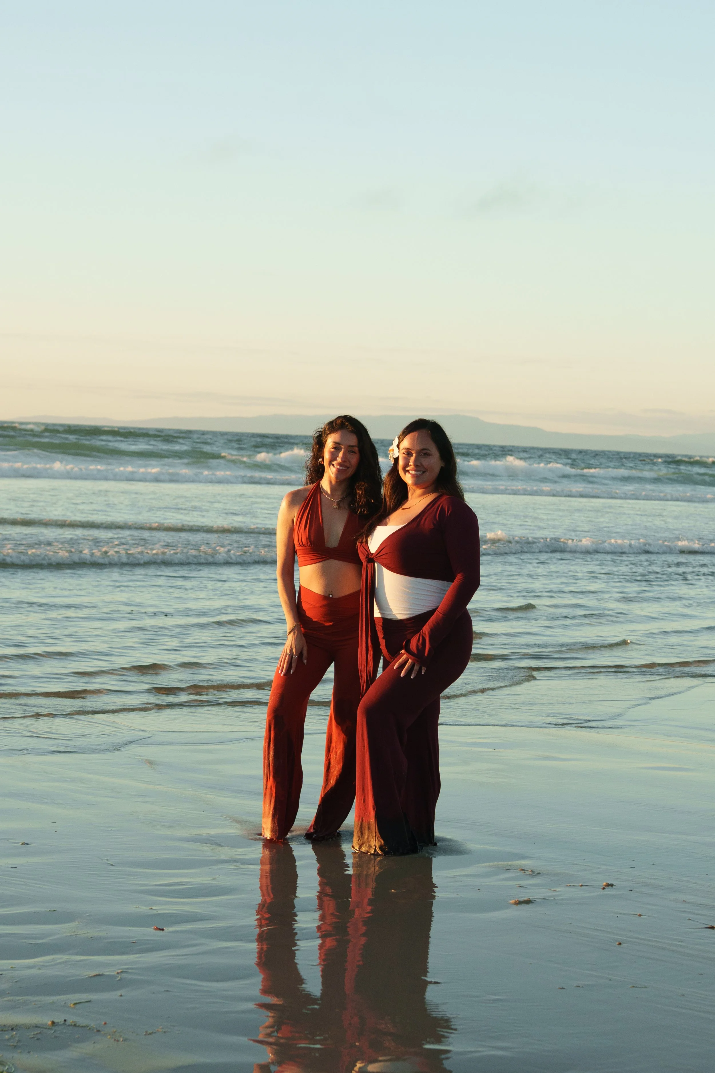 Two women standing in shallow ocean water at sunset, smiling at the camera, wearing matching rust-colored outfits, with a distant landmass in the background.