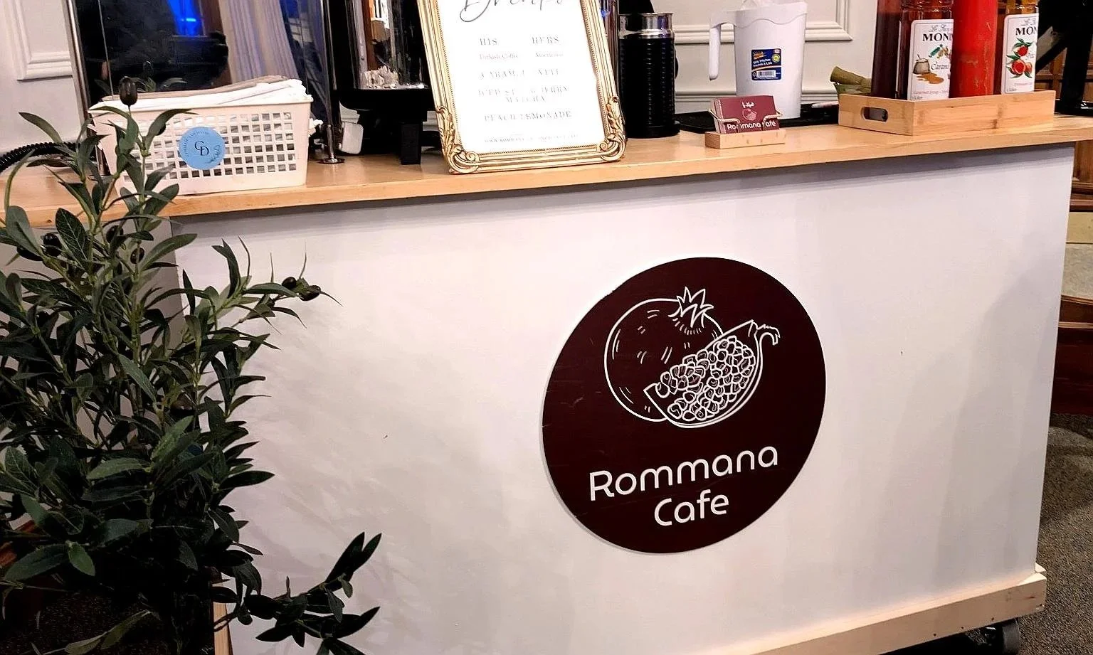 Coffee Cart with a round sign featuring a coffee cup and the cafe's name, coffee machines, and white basket on the countertop, with a small plant in the foreground.