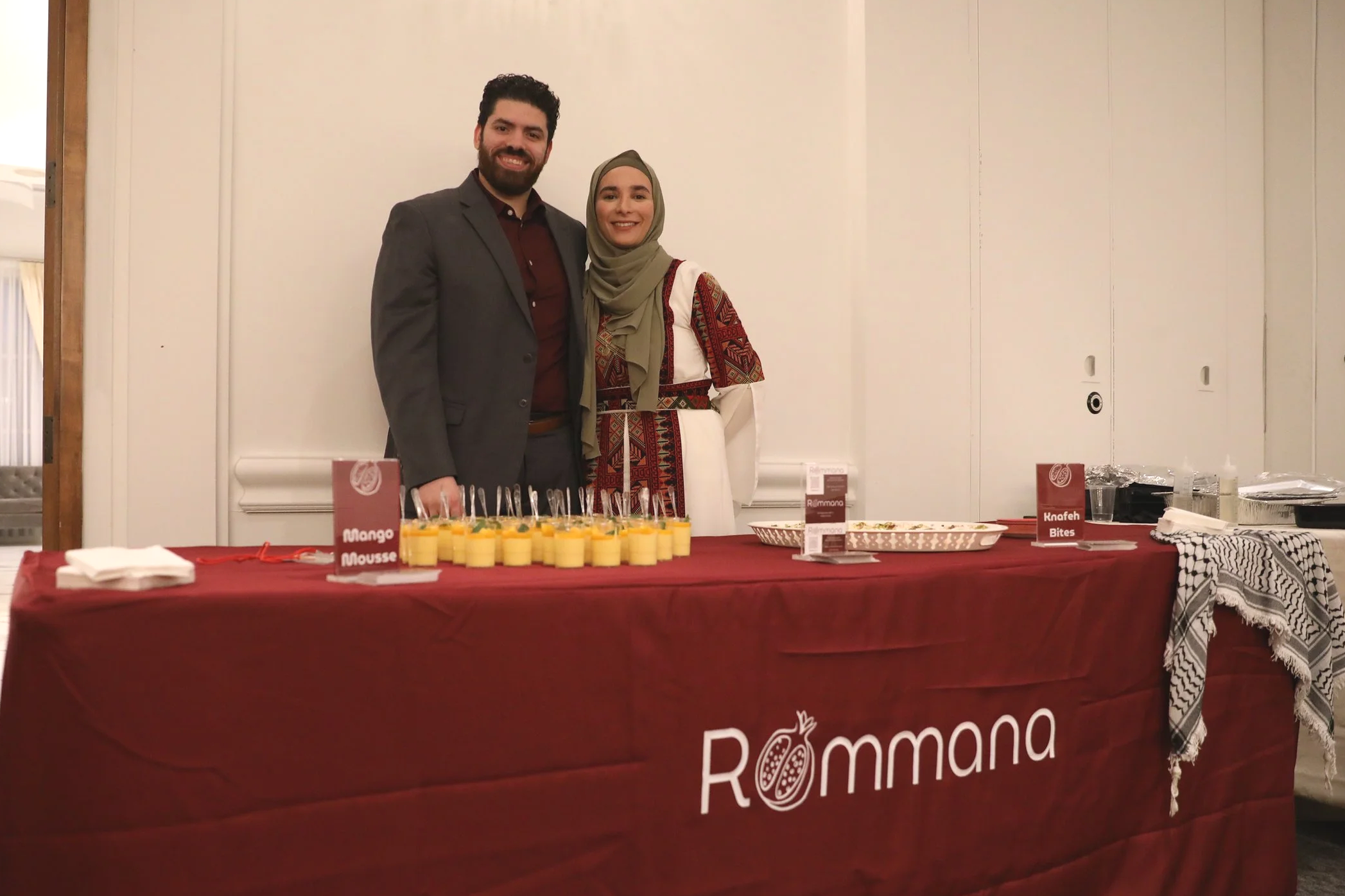 Two people standing behind a table covered with a red cloth that has 'Rmmana' written on it. The table displays small glasses of mango mousse. The man is wearing a dark suit with a maroon shirt, and the woman is wearing a traditional dress with a beige headscarf. They are smiling at the camera in a white-walled room.