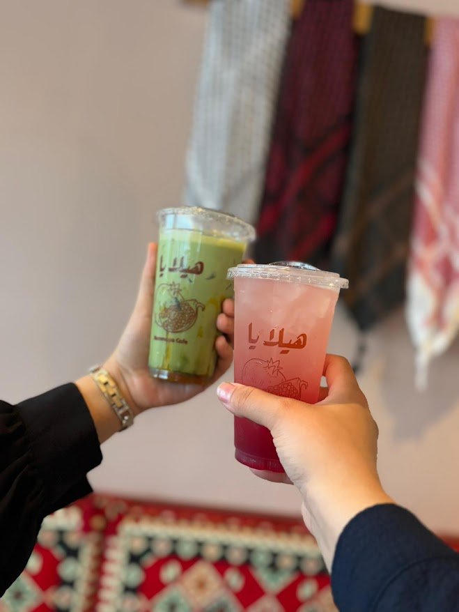 Two people holding colorful drinks in plastic cups with Arabic script, one green and one pinkish-red, in front of a display of fabrics.