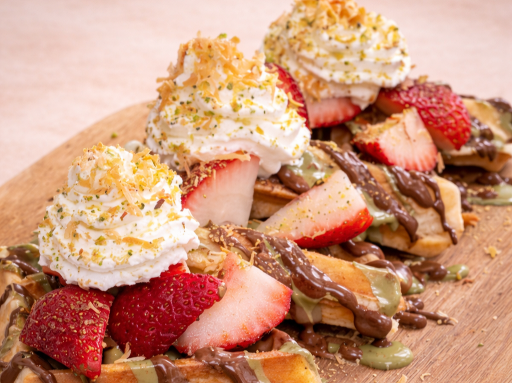 Belgian waffles topped with strawberries, whipped cream, chocolate drizzle, and crushed nuts on a wooden platter.