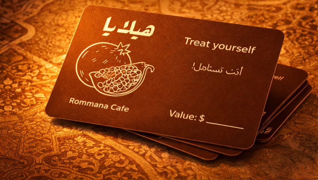 Business card for Rommana Cafe saying "Treat yourself" in English and Arabic, with an illustration of a bomb revealing a pile of food inside, placed on a patterned fabric surface.