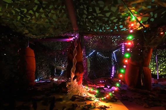A small sheltered outdoor area decorated with colorful string lights and camouflage fabric, creating a cozy, festive atmosphere.
