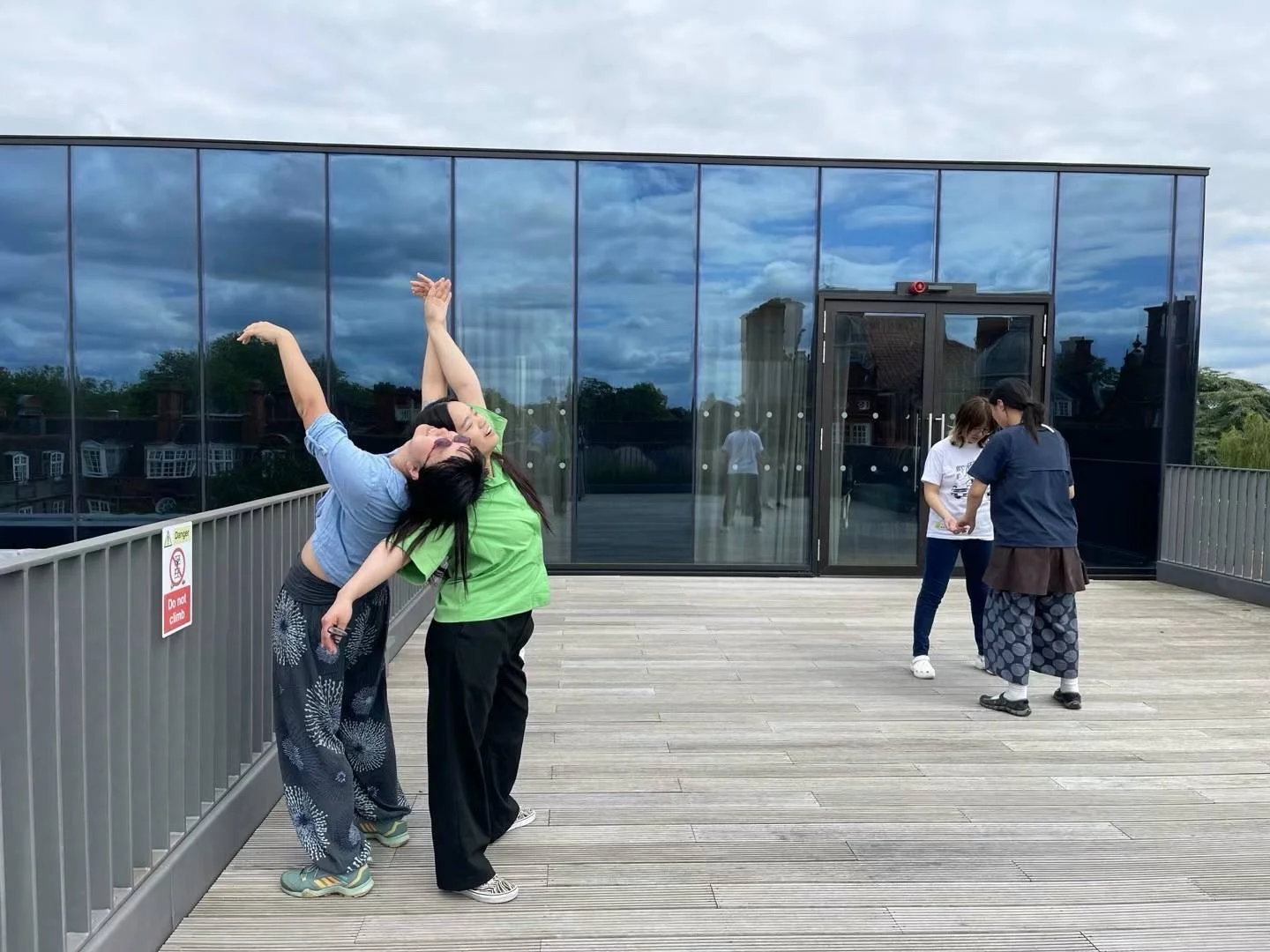 Two women are posing playfully on a rooftop with a modern building at the back, while two other women chat nearby. The sky is cloudy.