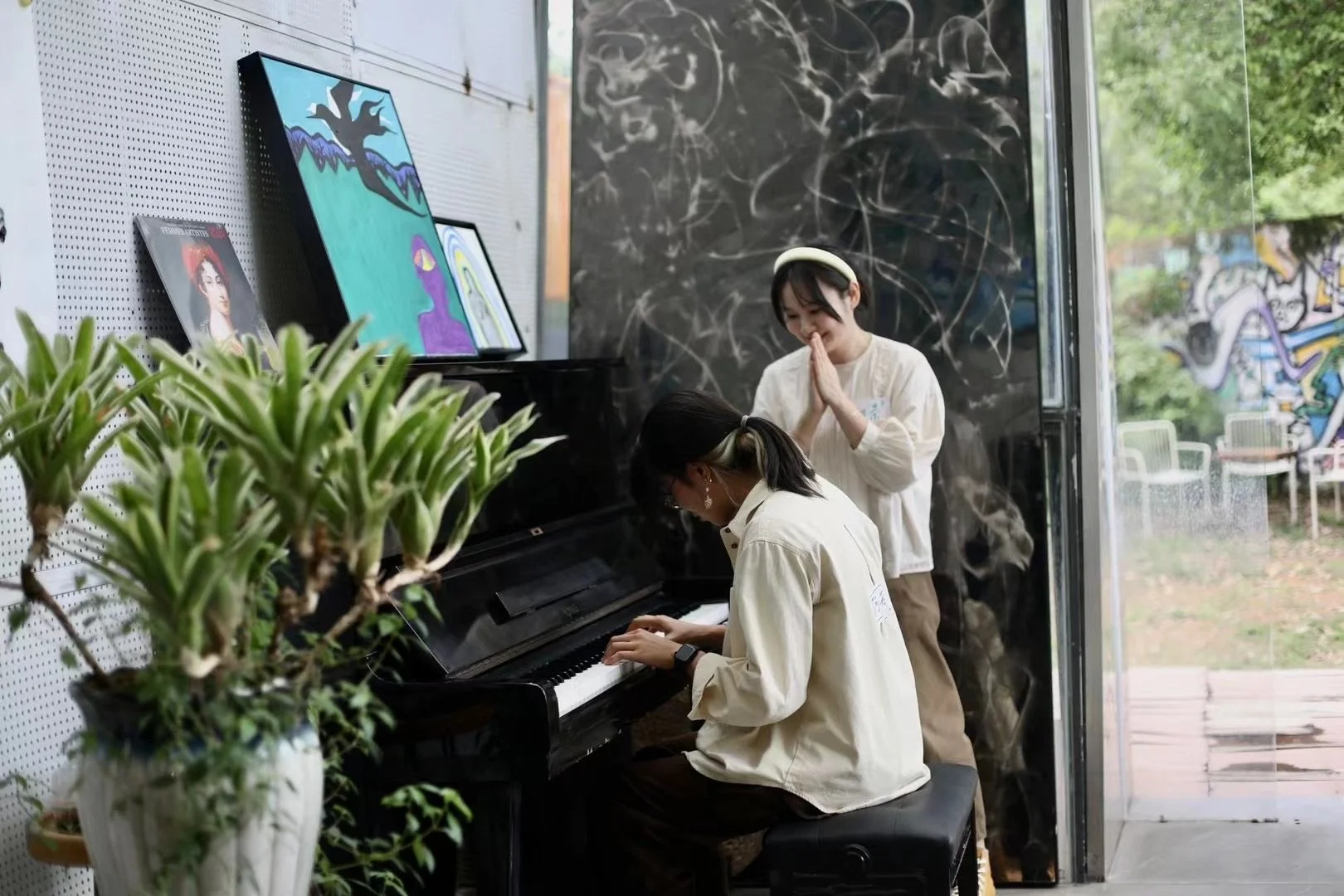 Two women inside a modern art gallery, one playing a black piano and the other with clasped hands watching attentively, with colorful paintings on the wall and plants nearby.