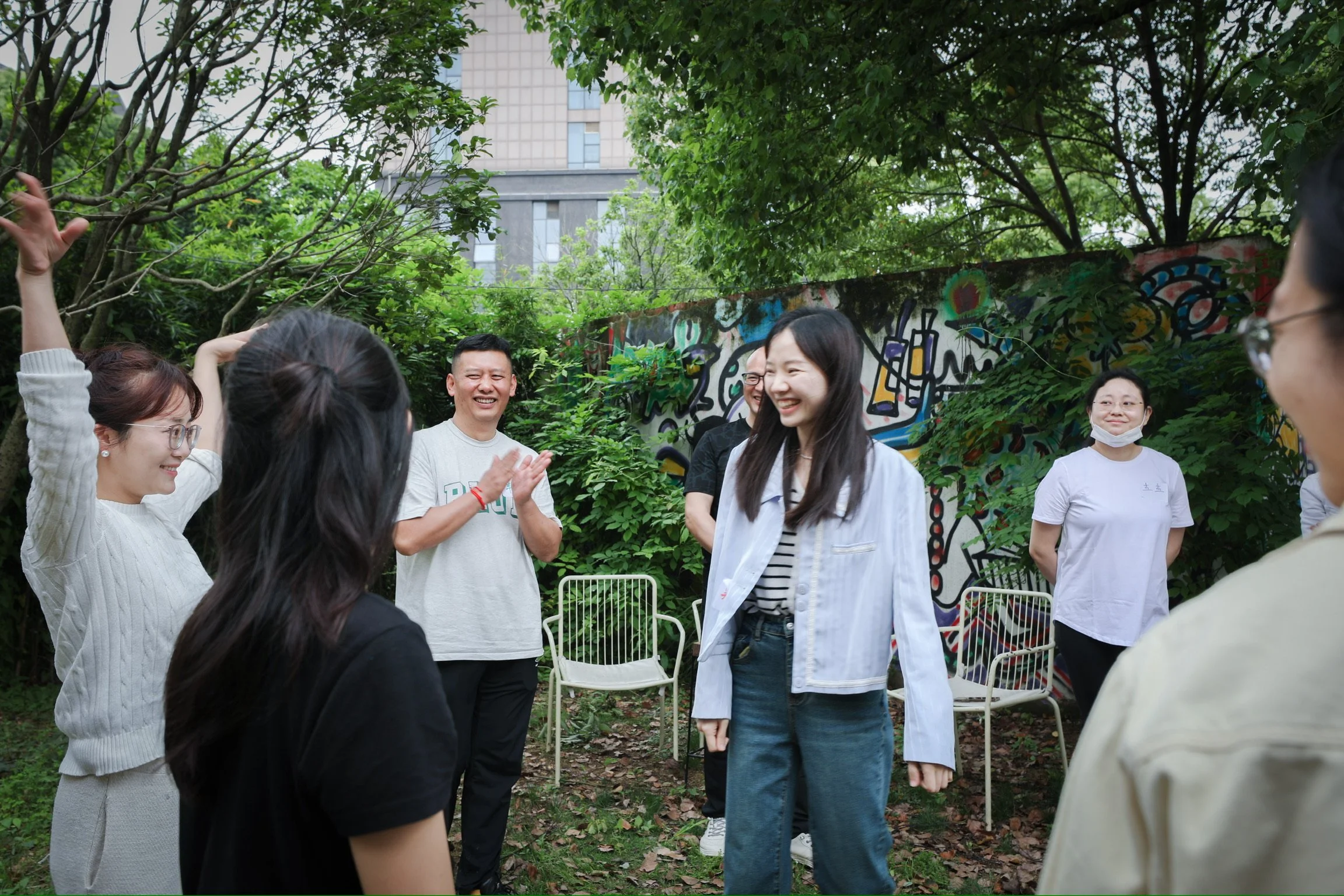 Group of people smiling and laughing outdoors in a green garden with trees and graffiti mural, some wearing masks
