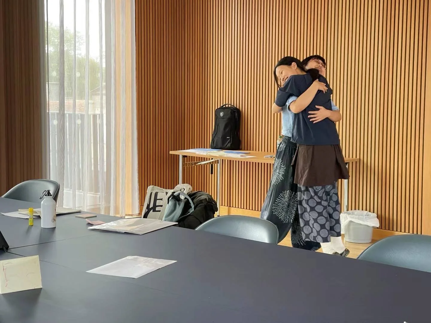 Two people embracing each other in a hug in a room with wooden wall paneling, a table with chairs, and personal items on the table and floor.