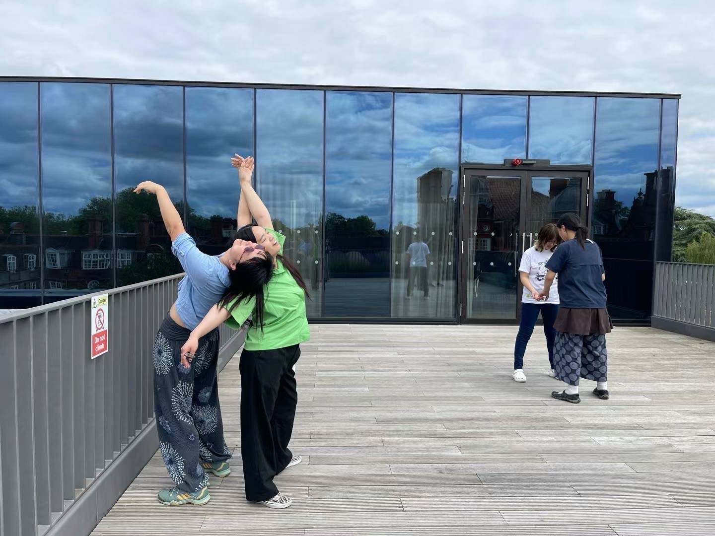 Four women dancing and talking on a rooftop terrace with glass walls, dark clouds in the sky, and a warning sign on the fence.