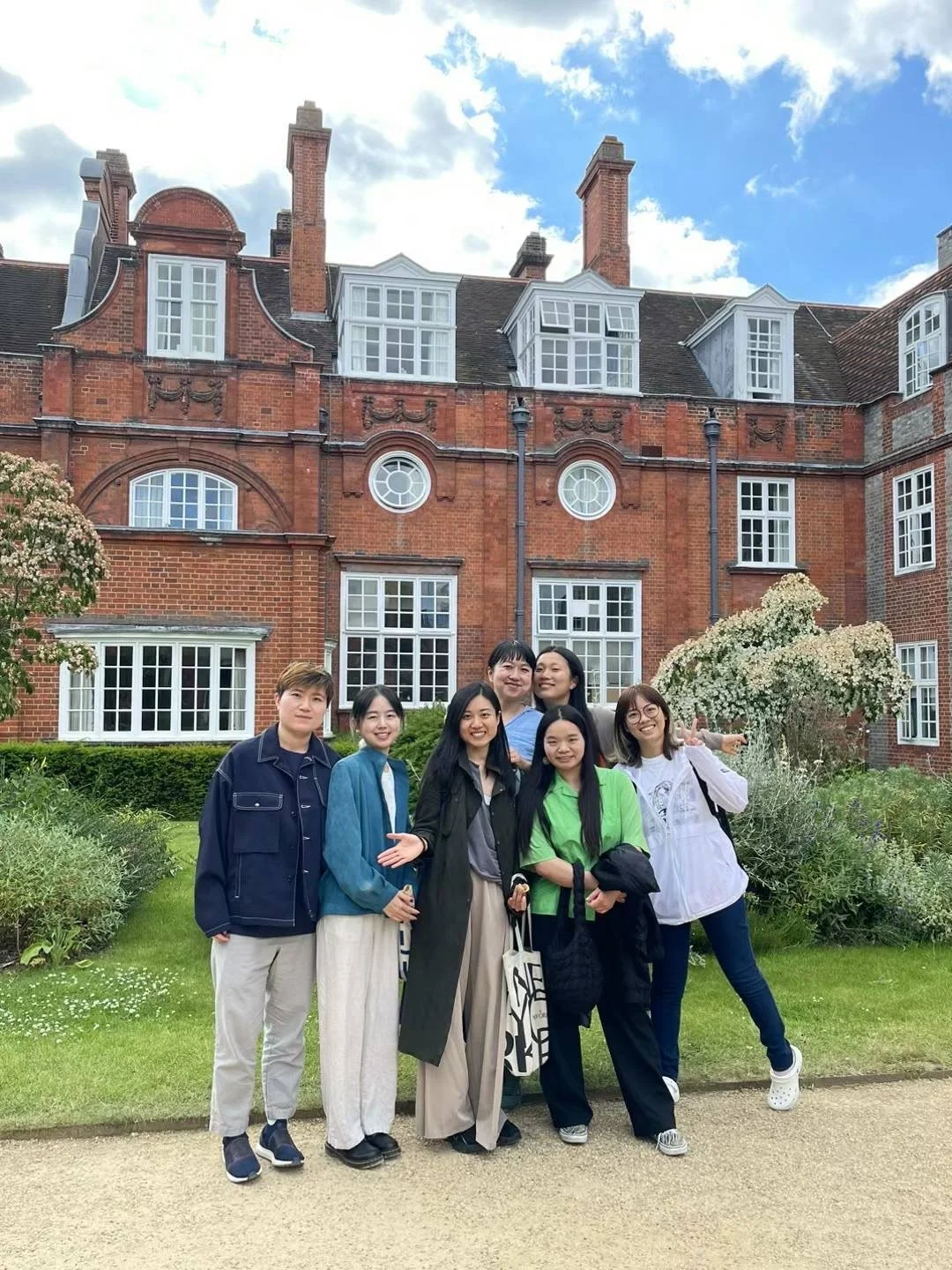 Group of six young women standing in front of a red brick historic building with large windows and a garden, during daytime with blue sky and clouds.