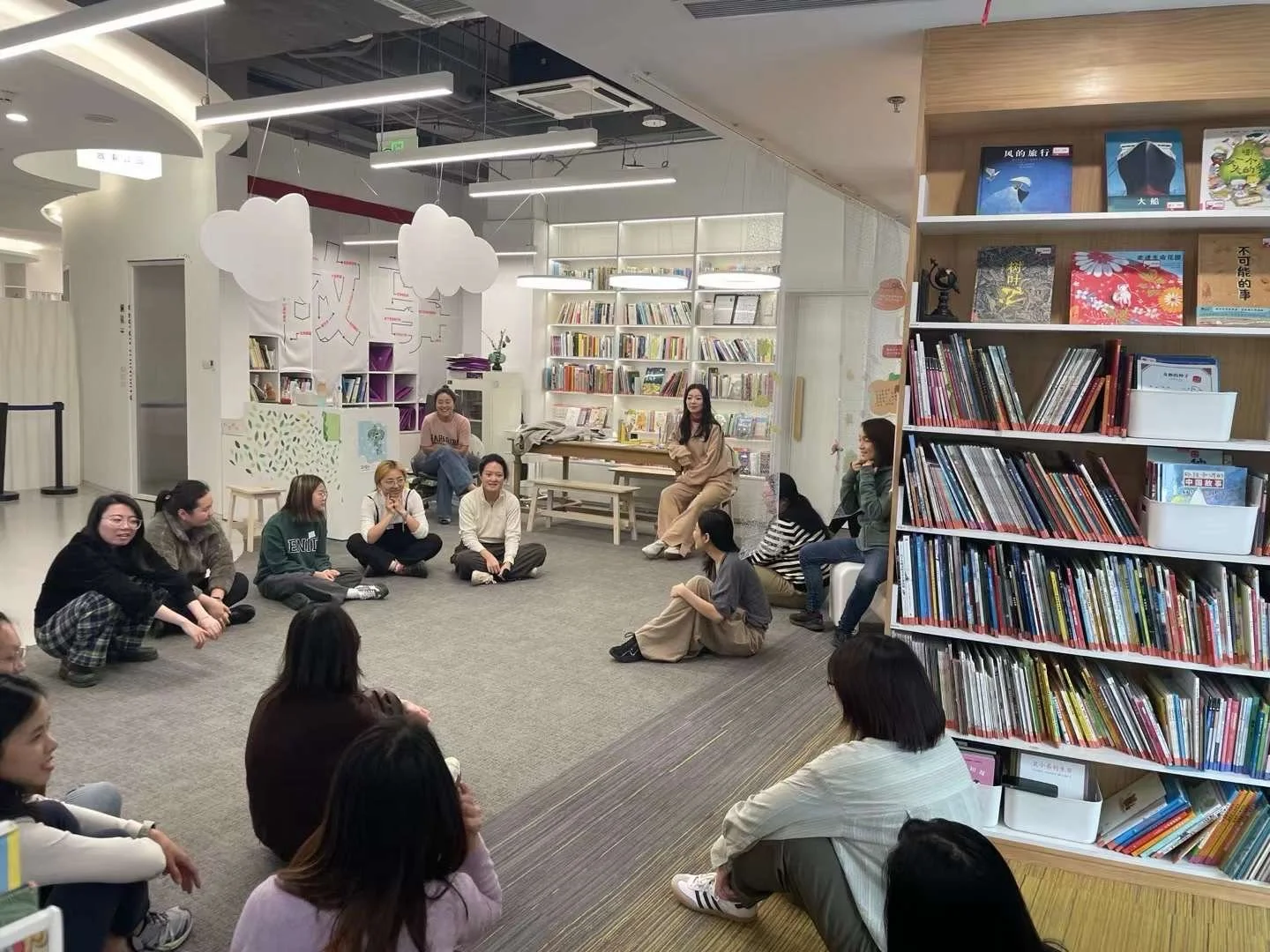 A group of women sitting on the floor and on chairs in a circle inside a library or bookstore, engaging in a discussion or activity, surrounded by bookshelves and modern decor.