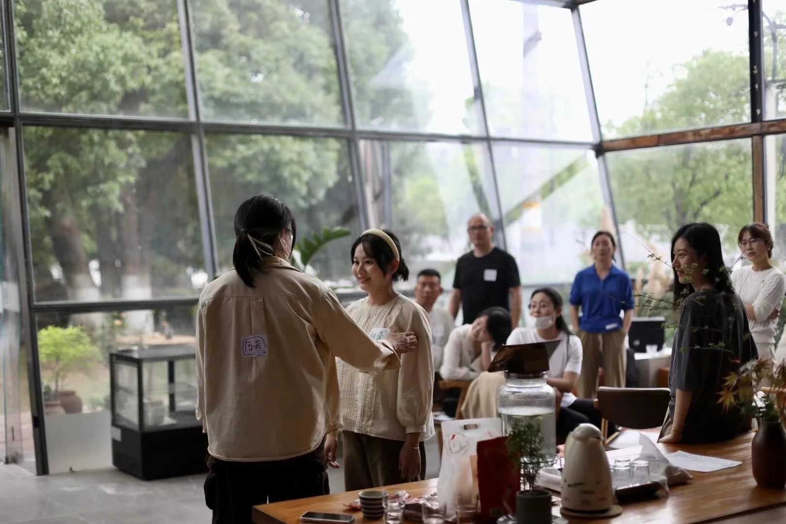 A woman in a beige jacket is pinning a name tag onto another woman’s cream-colored outfit in a spacious, glass-walled room filled with people.