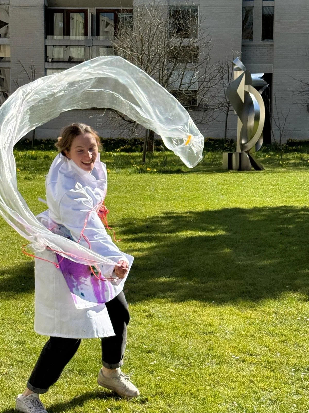 A woman outdoors on a sunny day with a plastic sheet flying above her, smiling and dressed in a white coat and black pants, with a modern sculpture and building in the background.