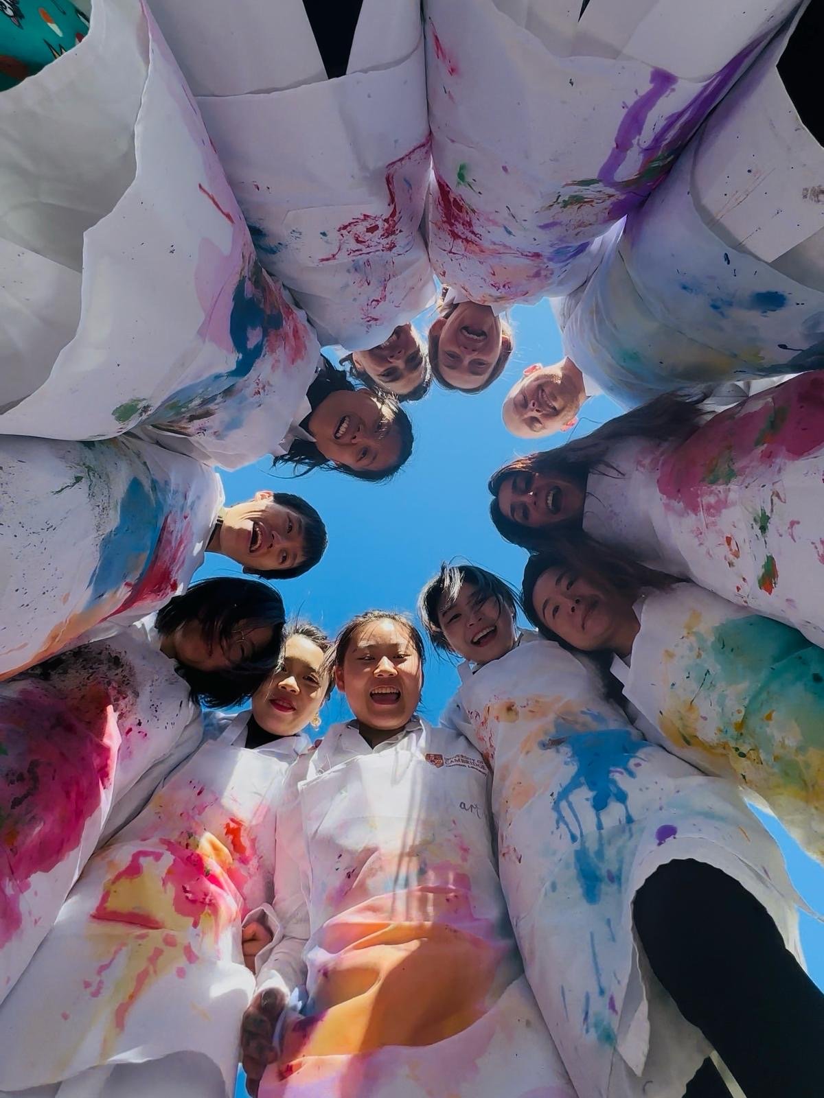 Group of people in white aprons covered in colorful paint, standing in a circle and looking down at the camera, smiling and laughing against a clear blue sky.