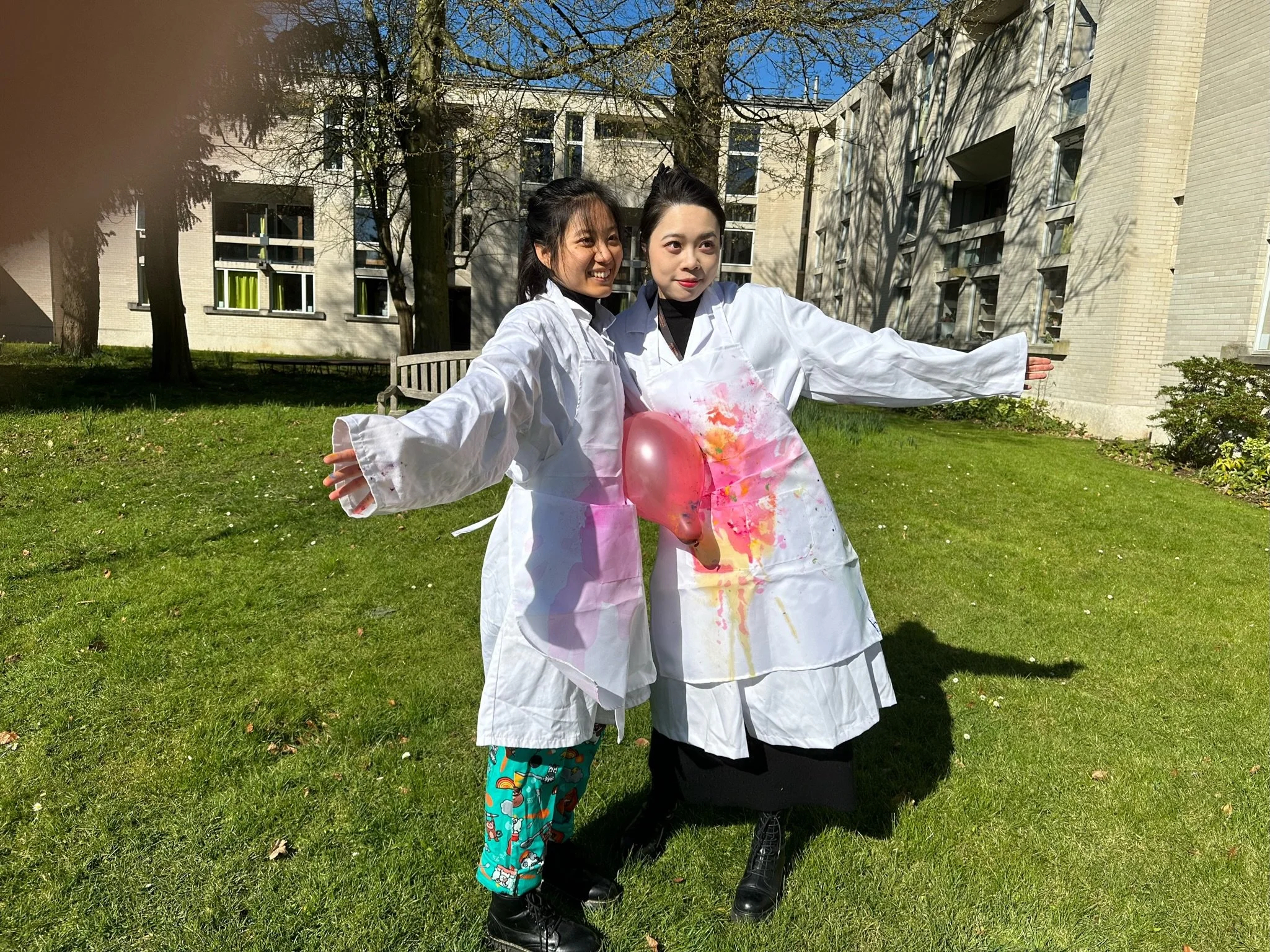 Two women in white smocks with pink paint splatters, one holding a pink balloon, standing outdoors on a grassy area with trees and a multi-story building in the background.