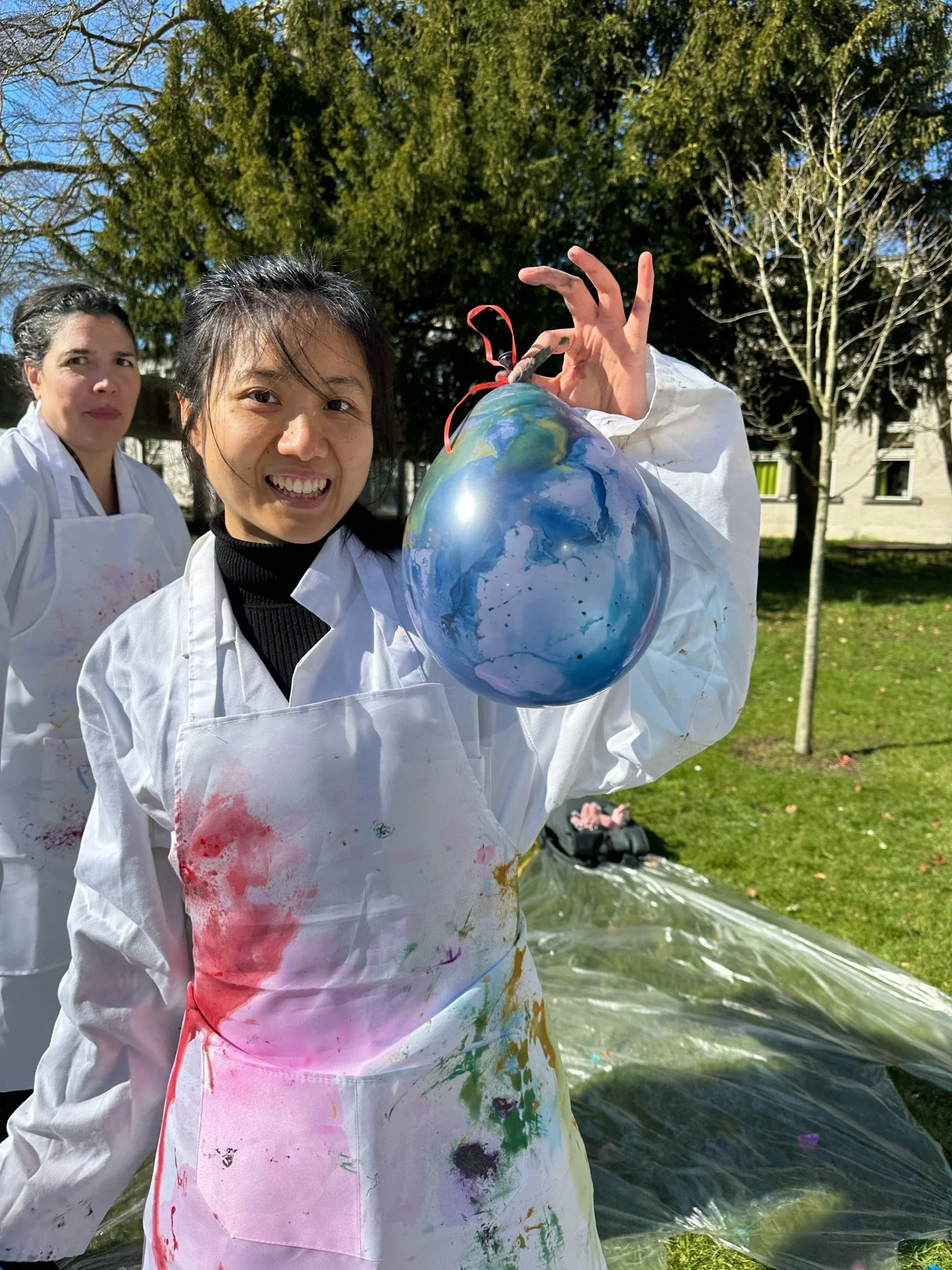 A woman wearing a white painter's apron with colorful paint stains holds up a painted globe with a red string. Two women in similar aprons are in the background outside on a sunny day with trees and a building behind them.