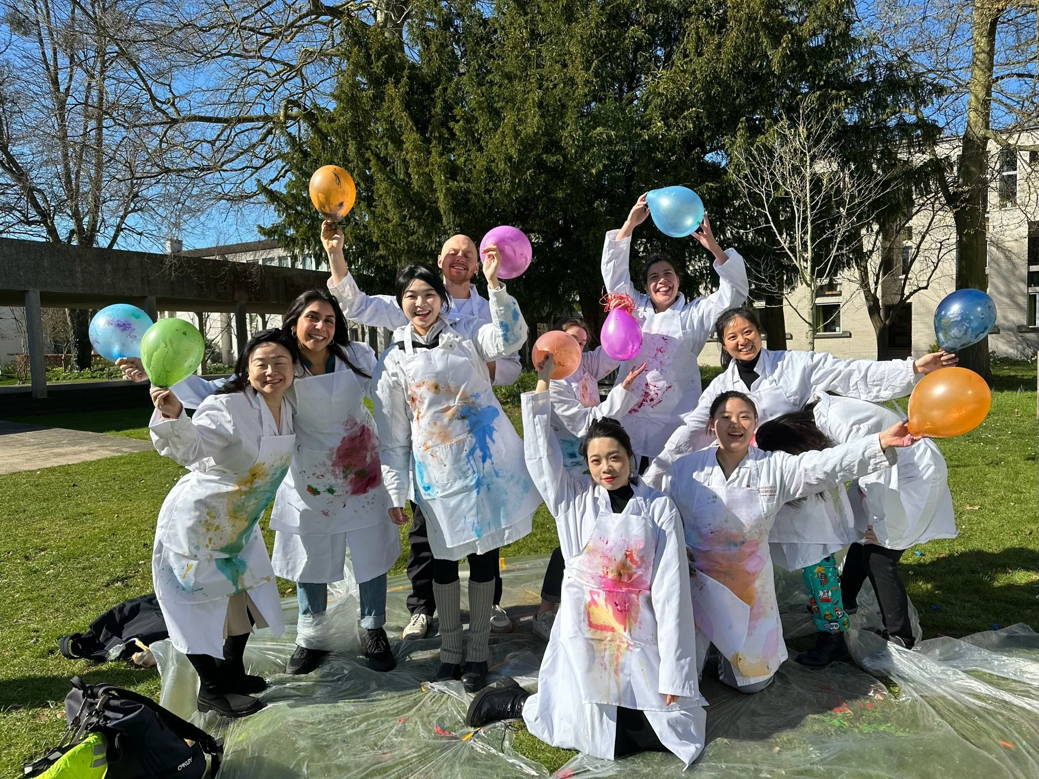 Group of people wearing white aprons with colorful paint splatters, holding and throwing colorful balloons outdoors on a sunny day with trees and building in the background.