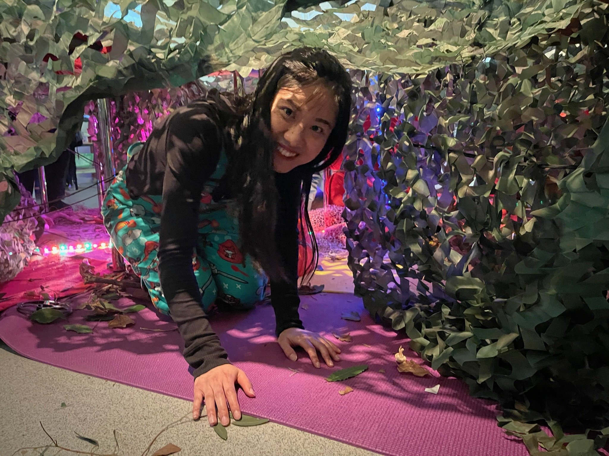 A woman crawling on all fours inside a space decorated with fake foliage, pink and purple lights, and leaves scattered on the pink yoga mat.