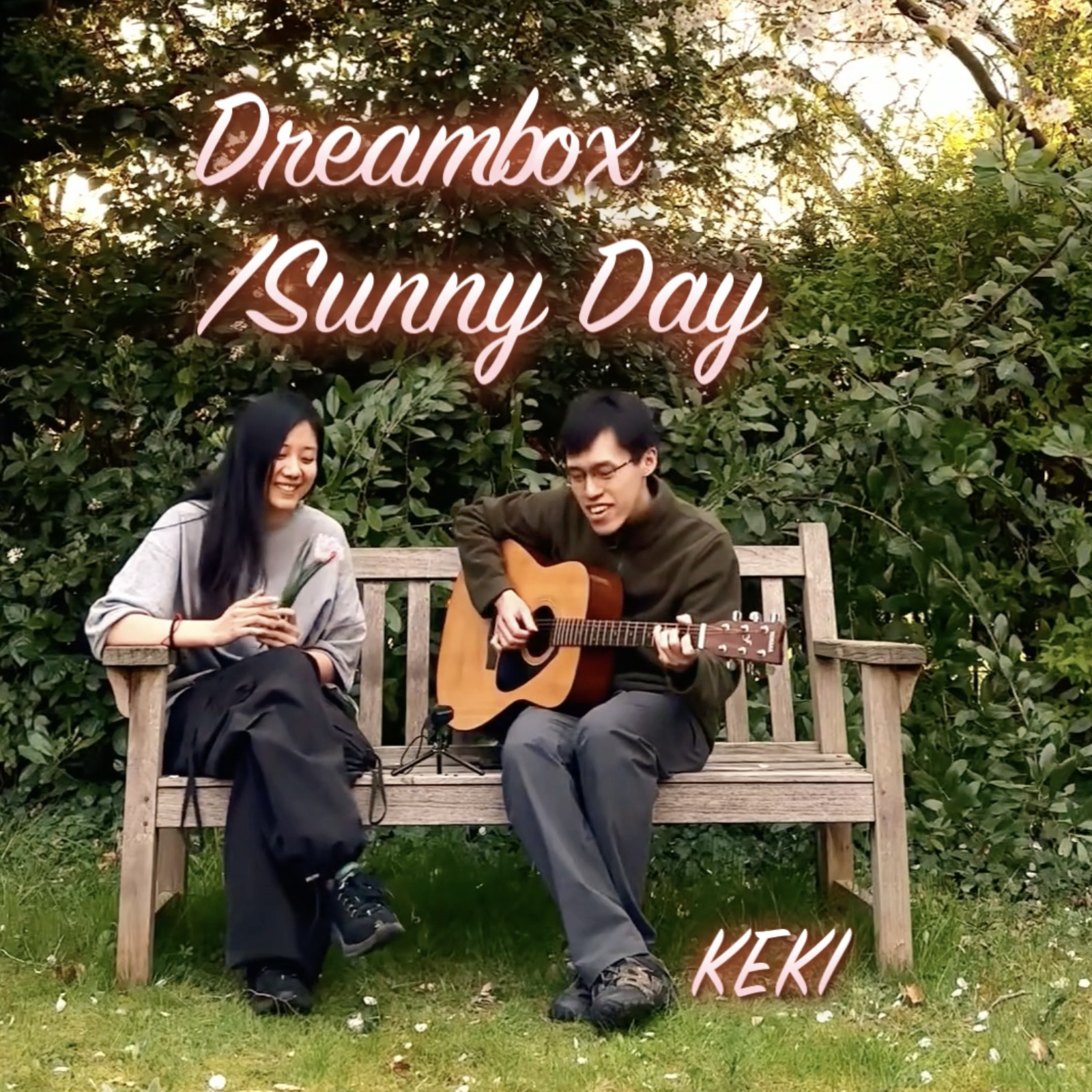 A man playing guitar and a woman holding a flower sitting on a wooden bench in a garden during daytime. The background includes green foliage and sunlight filtering through the trees. Overlaid text reads 'Dreambox / Sunny Day' and 'KEKI'.