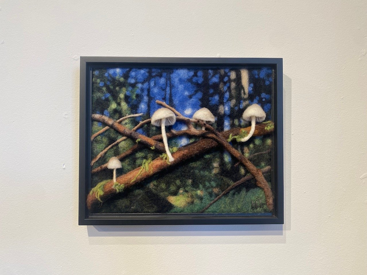 A framed felt art depicting a forest scene with a branch, mushrooms, and foliage, created using needle felting techniques.
