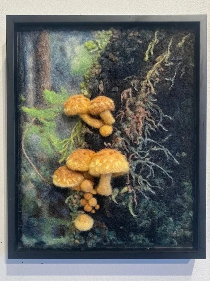 Felted artwork of yellow mushrooms and green foliage on a dark background, framed.