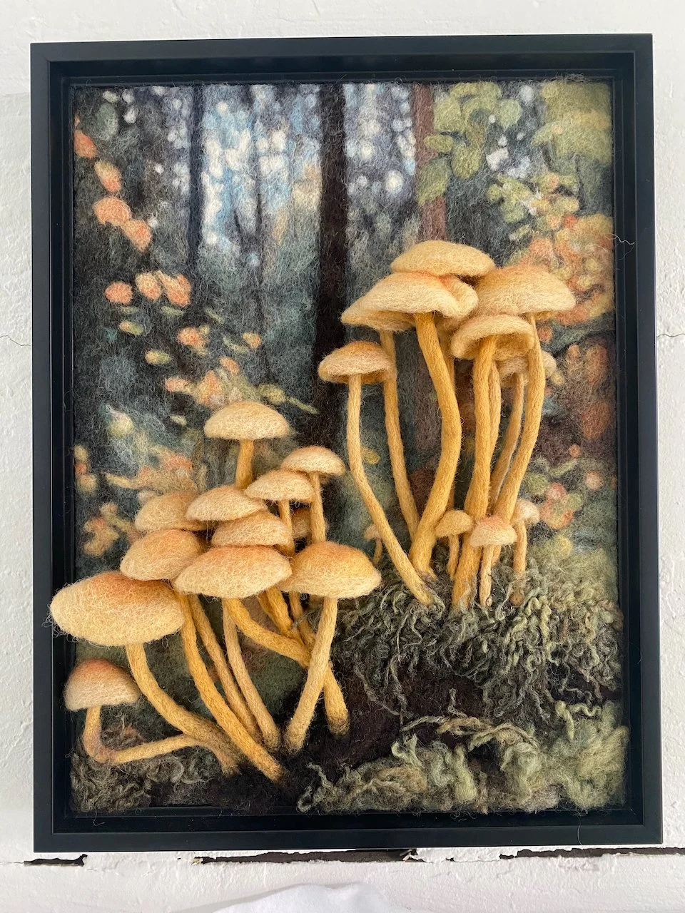 Felt artwork of a forest scene with cluster of mushrooms in the foreground, framed in black.