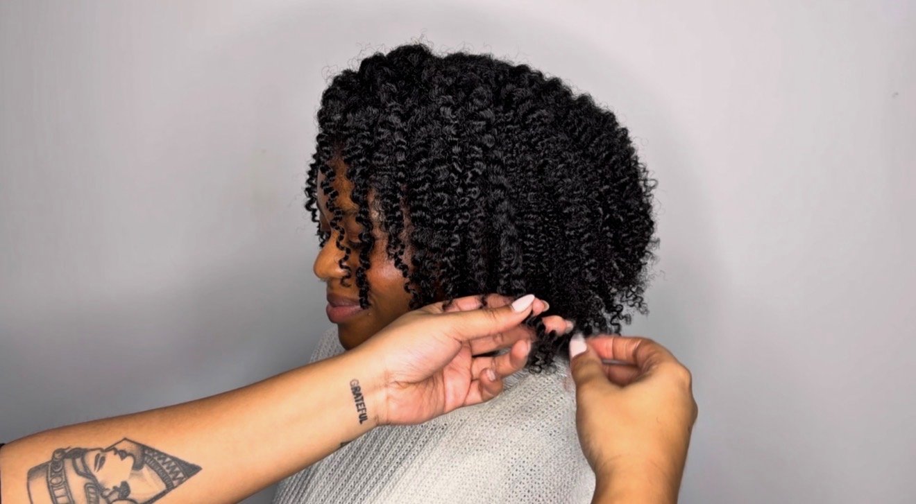 Une femme aux cheveux bouclés noirs est en train de faire des soins capillaires par une autre personne, qui touche ses cheveux.