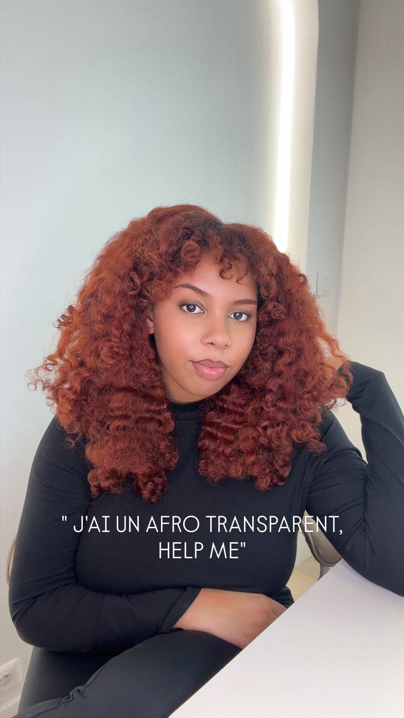 Une femme aux cheveux bouclés et roux, assise à une table, portant un haut noir, regardant l'appareil photo, avec une lumière naturelle en arrière-plan, texte en français indiquant qu'elle a un afro transparent et demande de l'aider.