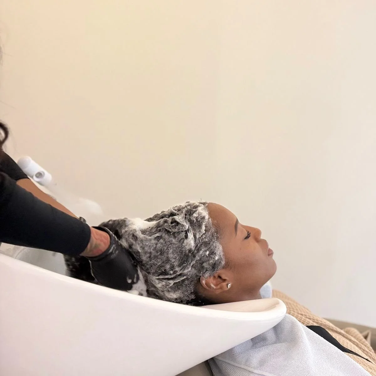 Une femme est en train de faire un shampooing dans un salon de coiffure, allongée avec la tête inclinée dans un bassin de lavage, tandis qu'une personne lave ses cheveux avec de la mousse.