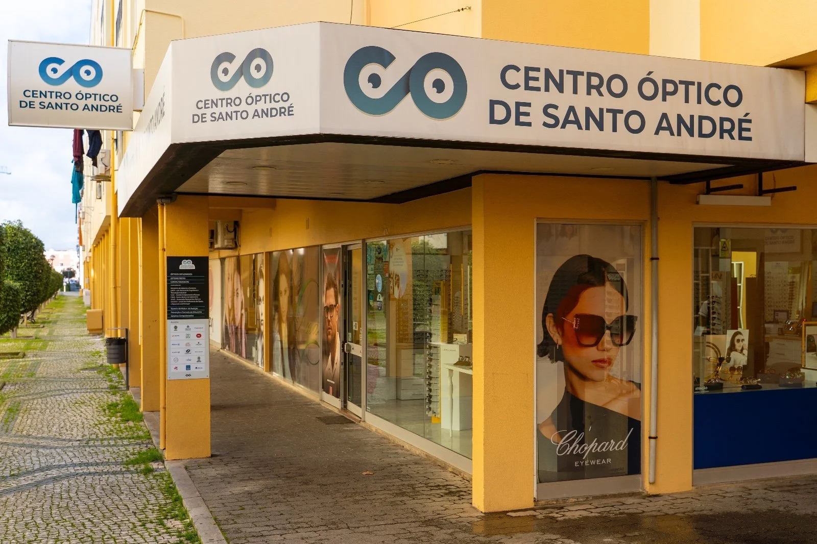 Exterior view of a yellow building with signs for 'Centro Óptico de Santo André' featuring an eyeglasses logo, and large posters of sunglasses displayed in the windows.