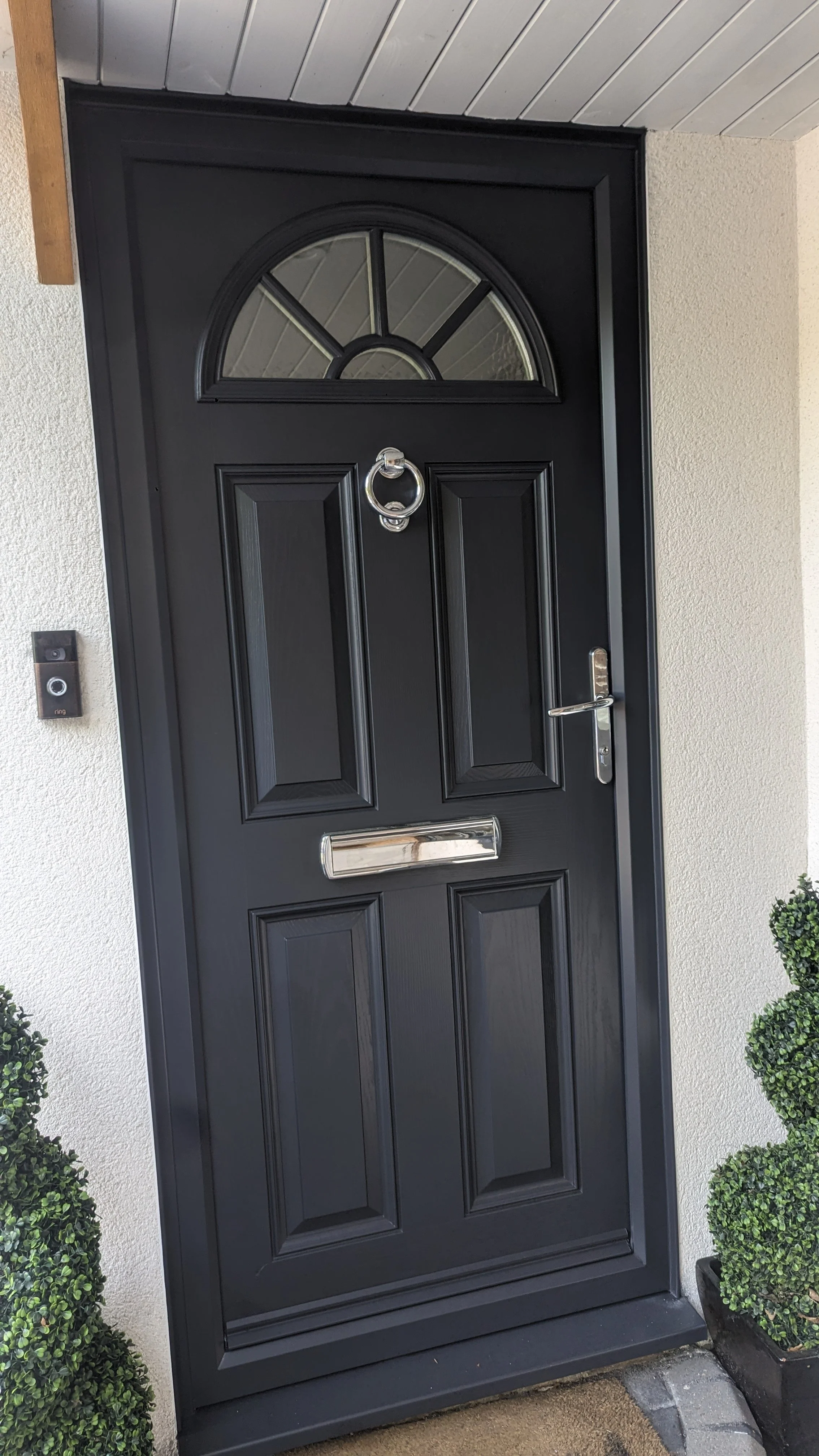 Black front door with a semi-circular window at the top, silver door handle, mail slot, round door knocker, keypad on the wall to the left, and potted bushes on both sides.