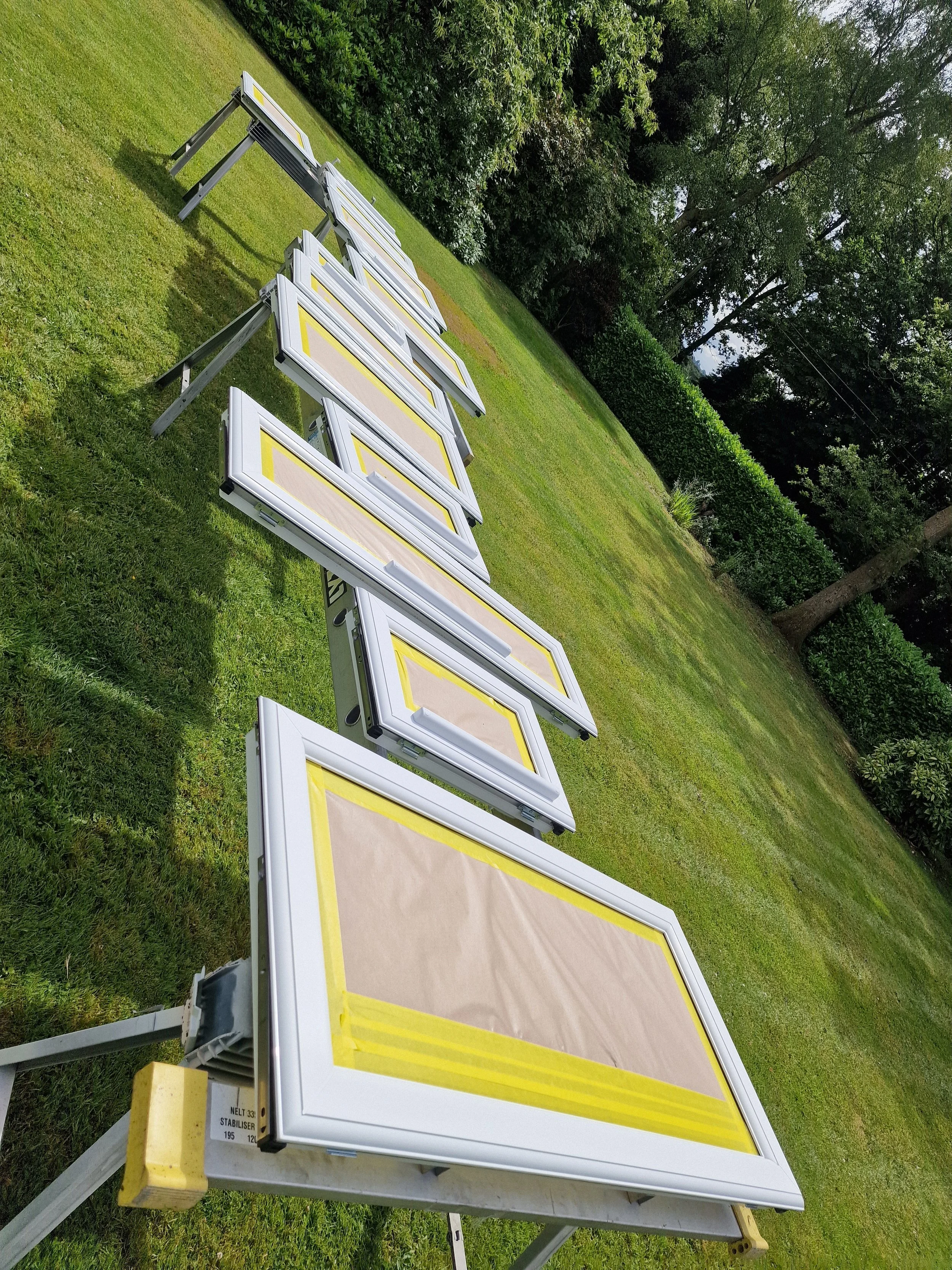 Multiple white and yellow frames with brown paper covers positioned on a grassy lawn, with trees and bushes in the background.