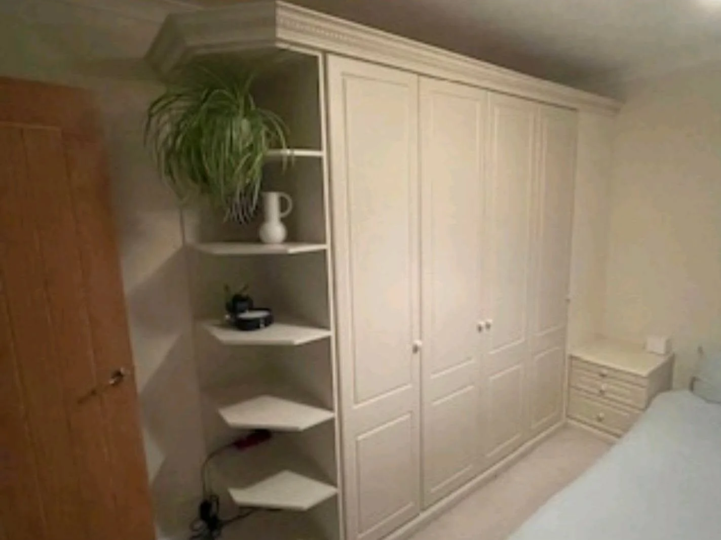 A bedroom with a large white wardrobe, a bedside table, a bed, and a corner shelf with a potted plant, a white vase, and other small items.