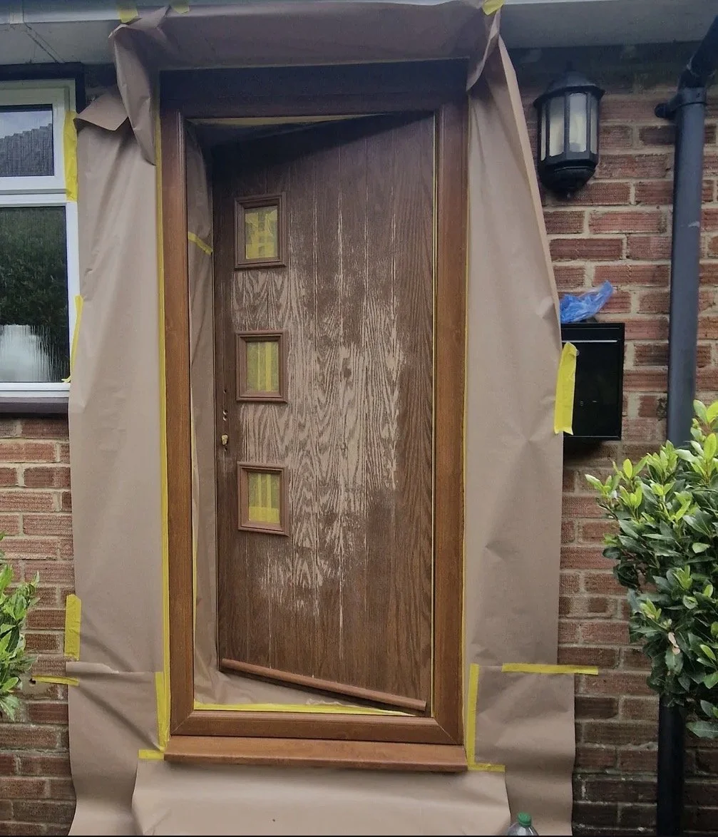 A wooden front door with three small windows, surrounded by beige paper and yellow painter's tape, mounted against a brick wall outside a house.