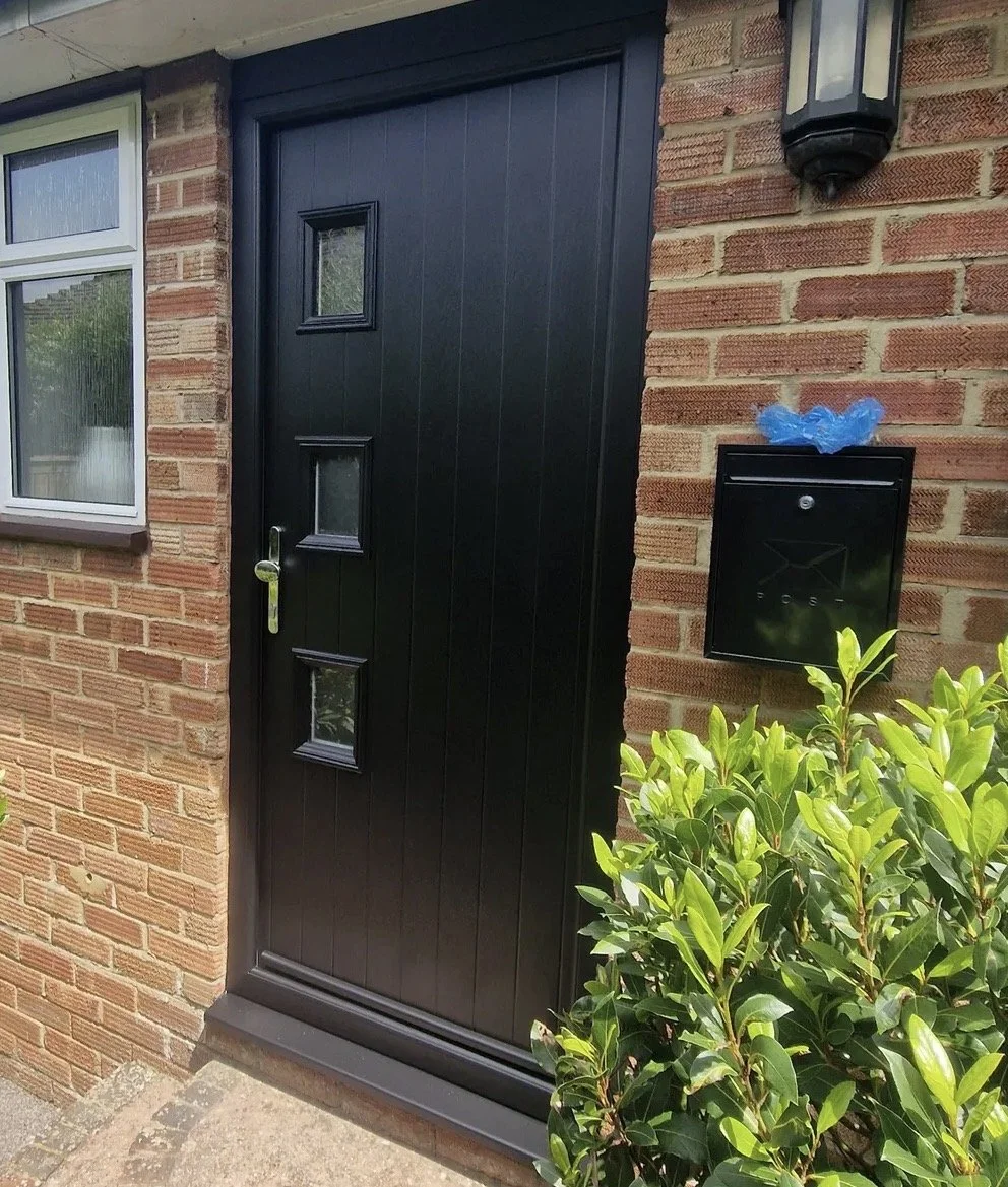 Black front door with three small square windows, a silver door handle, next to a brick wall with a light fixture and mail box with a blue plastic cover, surrounded by green bushes.