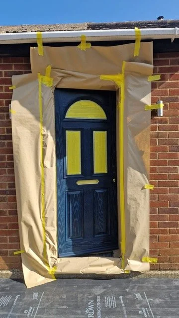Blue front door with a semi-circular window at the top, surrounded by brown paper and yellow painter's tape, on a brick house exterior.
