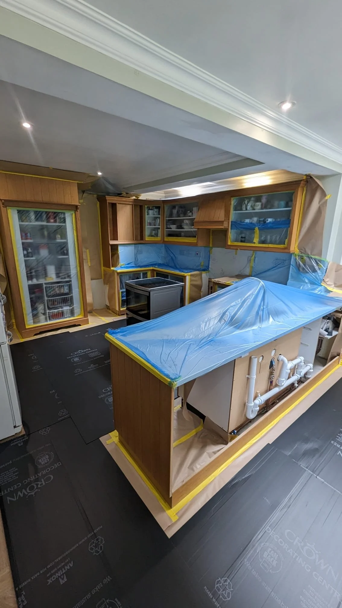 Kitchen under renovation, covered in blue protective plastic and masking tape, with cabinetry and appliances visible.