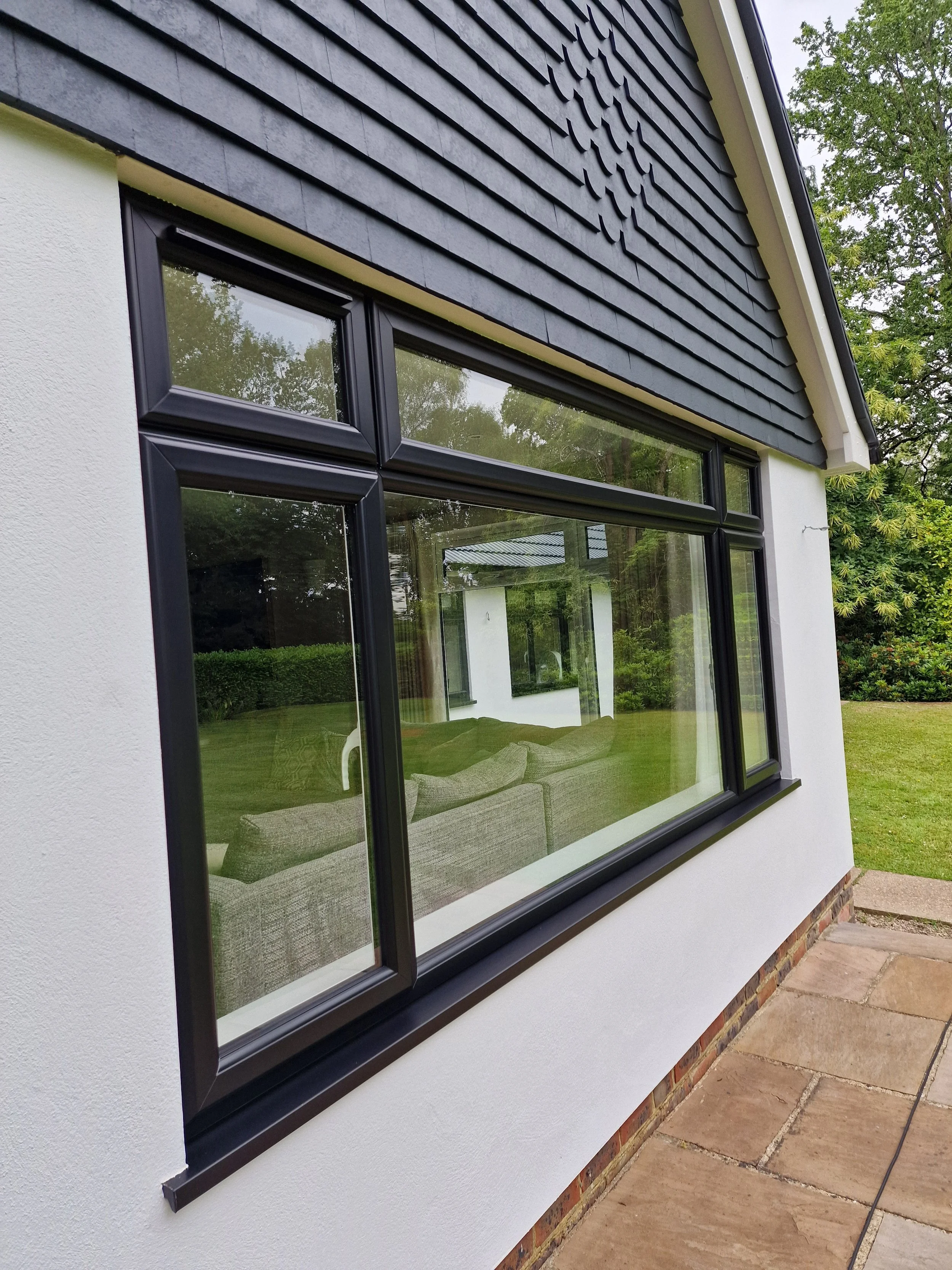 Close-up of modern black window frames on a white stucco wall, reflecting a grassy yard and white building, with gray shingle siding above and a paved stone patio outside.