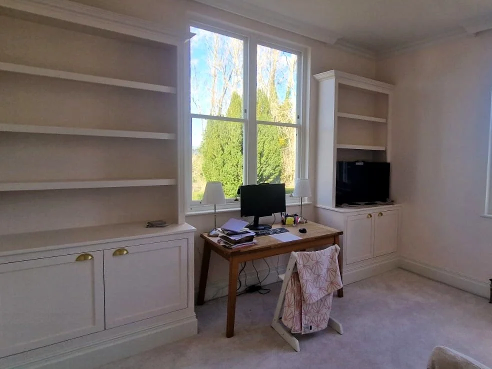 Home office with a wooden desk, computer, stacks of papers, and a chair with a floral towel, built-in white shelves, a TV, and large window showing green trees outside.